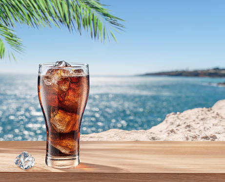 Glass of cola drink with ice cubes on table and blurred sparkling sea at the background.