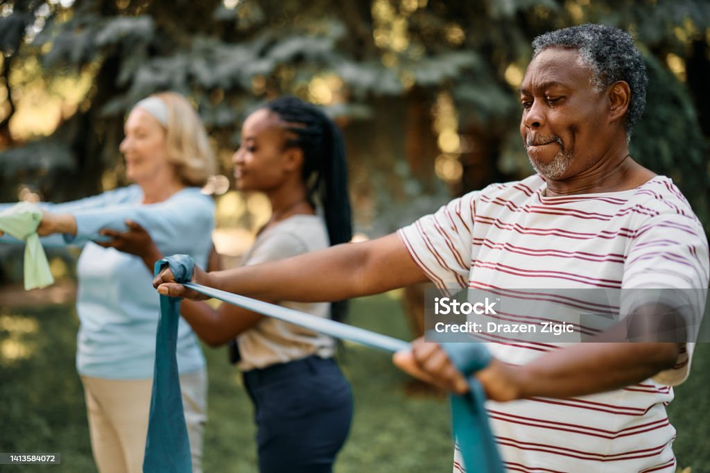 Black senior man using resistance band during exercise class in backyard of a nursing home. African American mature man practicing with power band on exercise class in nature. Exercising Stock Photo