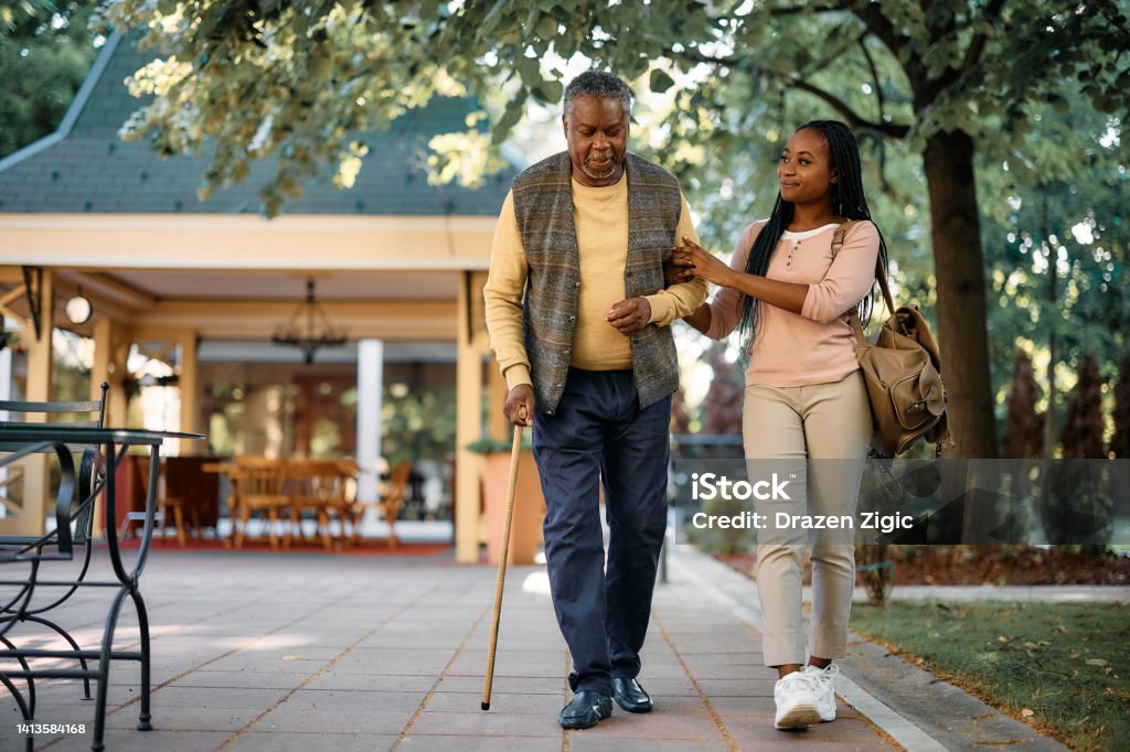 Black senior man with walking cane and his daughter taking a walk through the park of a nursing home. Smiling African American woman walking with her senior father while visiting him at nursing home. Senior Adult Stock Photo