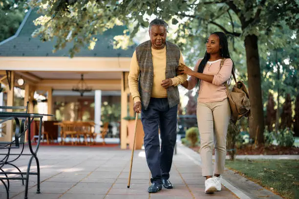 Photo of Black senior man with walking cane and his daughter taking a walk through the park of a nursing home.