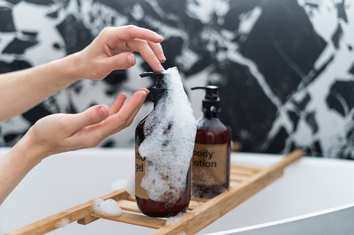 closeup of female hands use shower gel with foam for body care and cosmetics products on wooden tray on white ceramic bathtub in luxury bathroom with marble tile