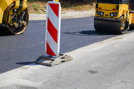 A red and white construction site indicator in front of road works laying new asphalt and steamroller.