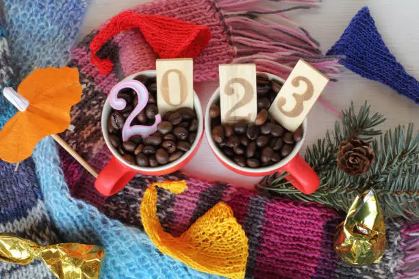 two cups filled with roasted coffee beans and the number 2023 on a table with colorful scarves and caps