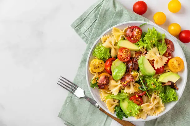 Cold summer pasta salad with bacon, tomatoes, avocado and mustard in a plate with fork on white background. Top view. Healthy diet food