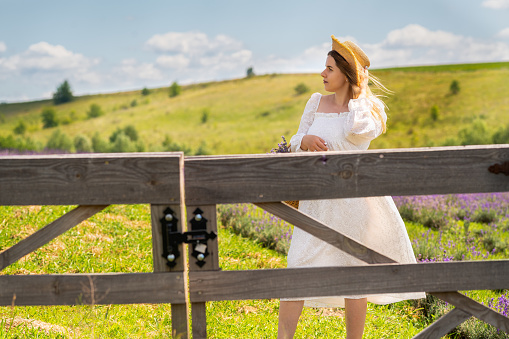 Young woman standing behind a closed rustic farm gate in fresh white cotton dress and straw sunhat looking off to the side