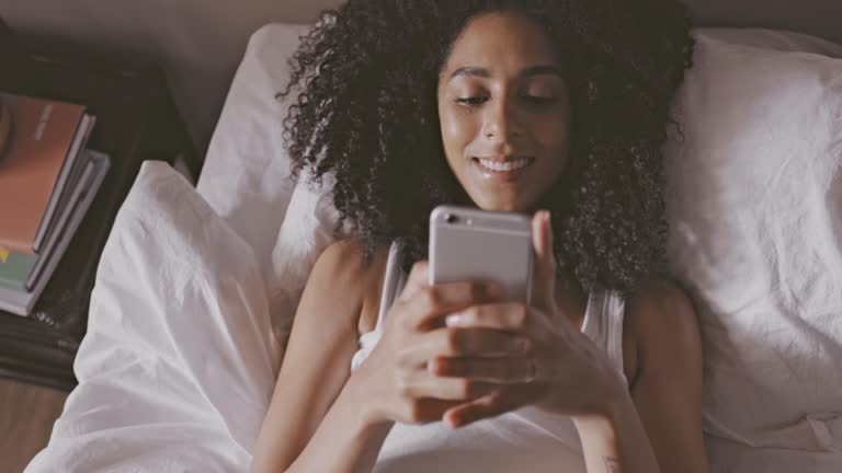 Happy black woman texting on a phone after waking up in the morning. Smiling African American female having an online conversation, replying to a dating app message while lying in bed at home