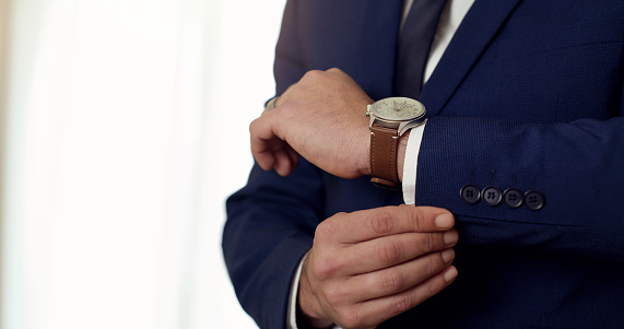 Elegant, formal and stylish man in a suit, buttoning his sleeve and wearing a watch while standing indoors. Closeup hands of businessman or groom looking neat and ready to leave for his wedding