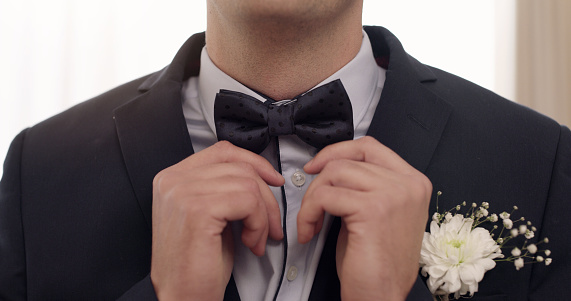 Wedding closeup of a man in a formal suit getting ready and preparing inside a bright room. Details of one bridal groom fixing or adjusting his bow tie while waiting for marriage on special occasion