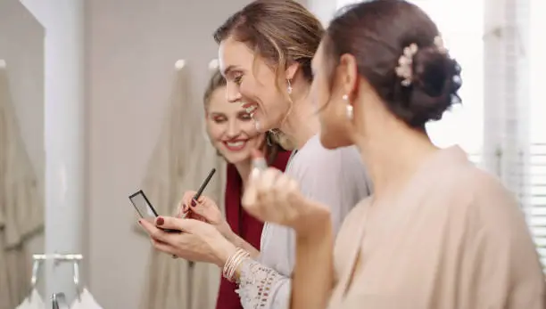 Group of happy, beautiful and elegant bridesmaid women getting ready, applying makeup and dressing in the bathroom for a wedding. Modern young and pretty ladies putting on cosmetics in a mirror