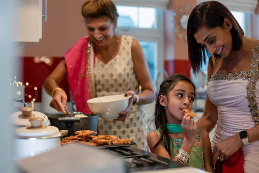 A close-up side view of a three gen female family cooking food for their family as they celebrate Diwali in the family home. The young girl is eating some fresh Jalebi from the baking tray and trying it as her grandmother serves the fresh food into serving dishes.