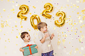 Happy smiling Kids are enjoying confetti near white wall with gold balloons. Celebrating New Year 2023. Christmas concept