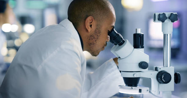 científico masculino enfocado que mira bajo el microscopio hace análisis de muestra de prueba de una vacuna de virus en un laboratorio de investigación científica. joven ingeniero genético negro especialista que trabaja con equipos futuristas - scientist lab coat doctor male fotografías e imágenes de stock