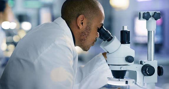 Focused male scientist looking under microscope does analysis of test sample of a virus vaccine in a scientific research lab. Young black genetic engineer specialist working with futuristic equipment