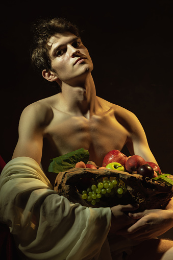 Creative remake of painting boy with a basket of fruit. Young handsome man over dark vintage background. Italian baroque style, art, creativity, vintage, comparison of eras concept.