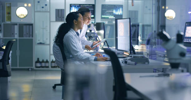 focused, serious medical scientists analyzing research scans on a computer, working late in the laboratory. lab workers examine and talk about results from a checkup while working overtime - estilo de vida imagens e fotografias de stock
