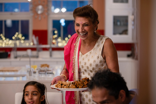 A selective focus front view of a grandmother bringing her freshly cooked onion bhajis and samosas and spring rolls to her impressed granddaughter who is licking her lips as well as the rest of her family. She has a cheerful proud smile on her face.