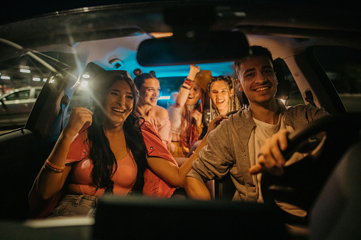 Group of young male and female friends riding in a car in city at night, man is driving the car.