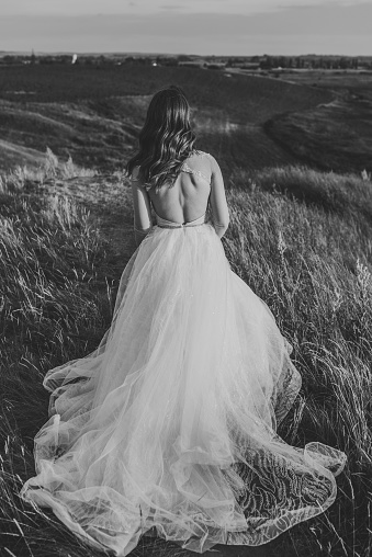 Bride holding her wedding dress and running in a field on the road. Woman in the mountains top. Girl walking on meadow in grass. Back view. Black and white photo.