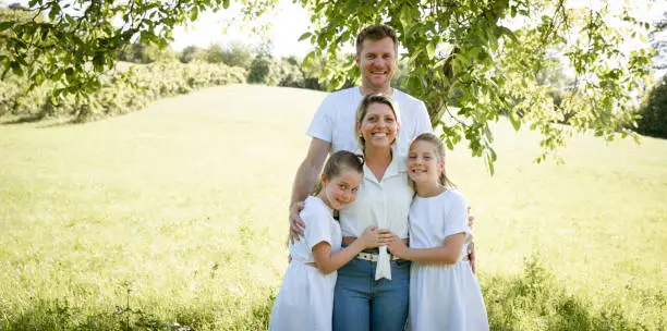 Photo of pretty family with two girls is standing in meadow near walnut tree and has light outfit on and jeans