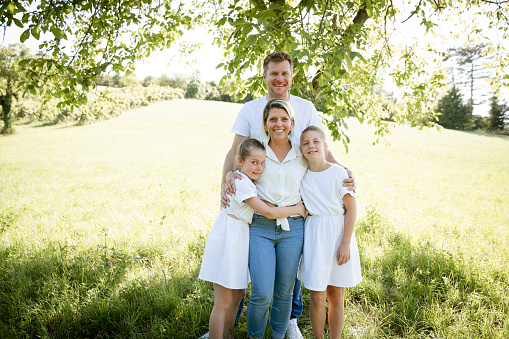 beautiful family with two girls is standing in meadow near walnut tree and has light outfit on and jeans and is happy