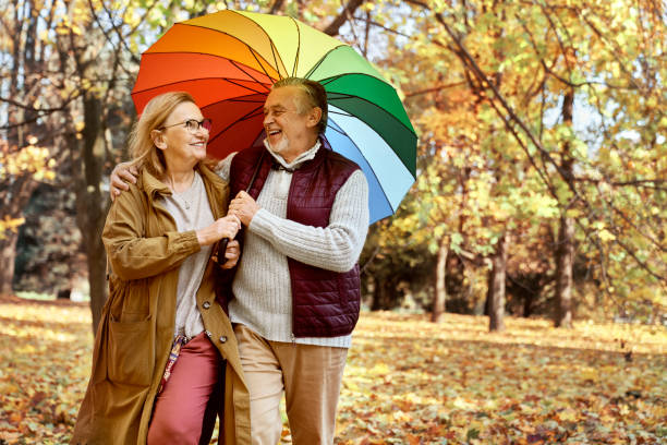 senior couple walking with umbrella at the park during the autumn - umbrella senior adult couple autumn imagens e fotografias de stock