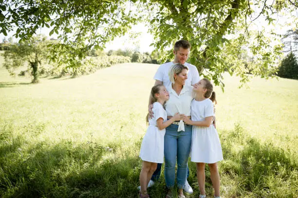 Photo of pretty family with two girls is standing in meadow near walnut tree and has light outfit on and jeans