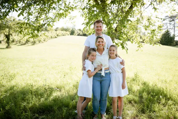 Photo of pretty family with two girls is standing in meadow near walnut tree and has light outfit on and jeans