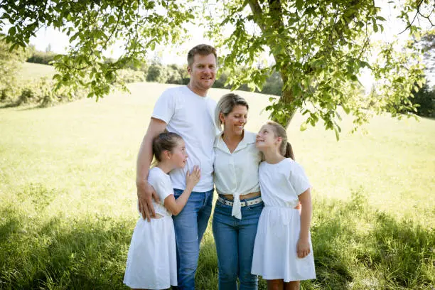 Photo of pretty family with two girls is standing in meadow near walnut tree and has light outfit on and jeans