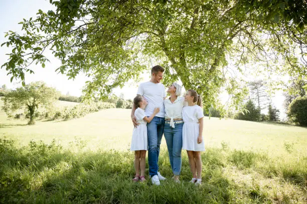 Photo of pretty family with two girls is standing in meadow near walnut tree and has light outfit on and jeans