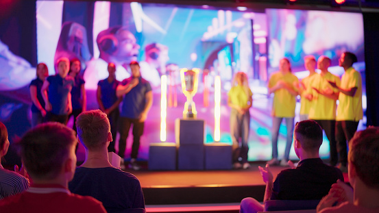 Diverse gamer group wearing yellow t-shirts with african ethnicity players and women on stage receiving a trophy. Audience in background