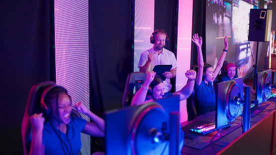 Diverse gamer group with african ethnicity players with a coach playing competitive computer game on a stage. Wearing headset and talking into the microphone to communicate with the blue pro team.
