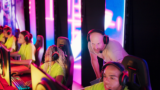 Diverse gamer group with african ethnicity players with a coach playing competitive computer game on a stage. Wearing headset and talking into the microphone to communicate with the yellow pro team.