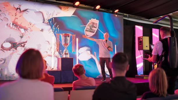 istock Esport commentator introducing competing teams. Cameraman filming, audience in background 1413616760