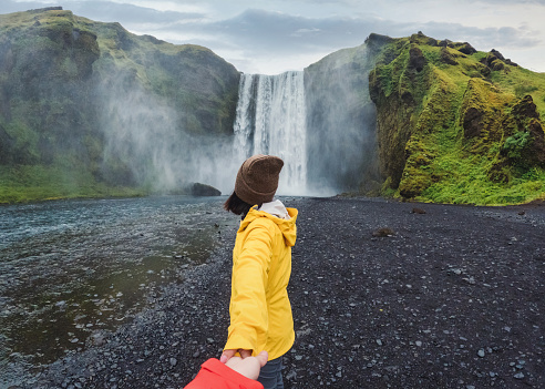 Asian woman in yellow jacket holding hands with couple at front of SkÃ³gafoss waterfall flowing on cliff in summer at South of Iceland