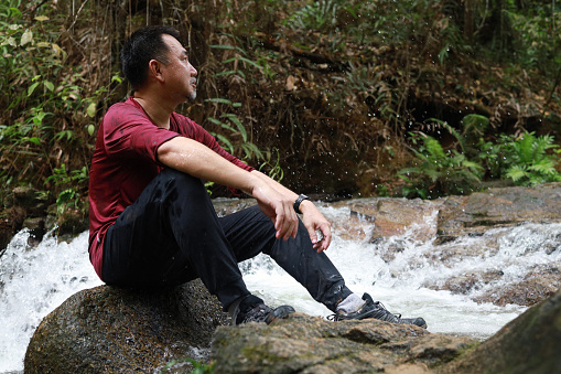 Portrait of an Asian man sitting on a rock and enjoying view of a waterfall in a tropical rainforest