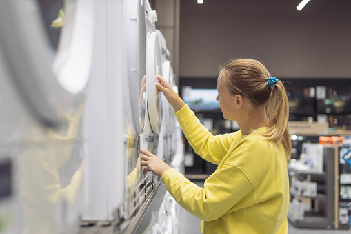 a fair-looking girl with blond hair tied in a ponytail in a yellow sweater, looking at a washing machine in a store. Close-up of her face and hands. High quality photo