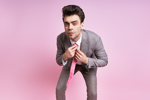 Confident man in full suit adjusting his necktie while standing against pink background