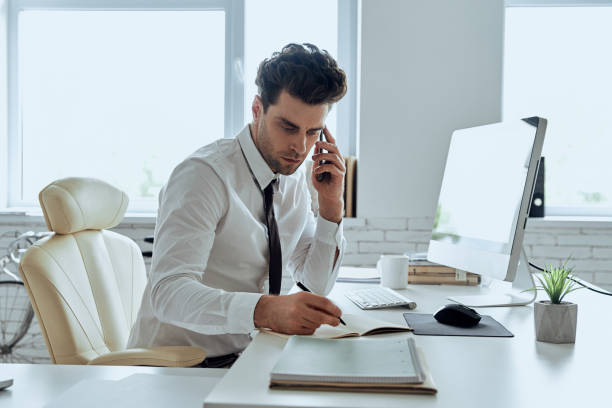 istock Confident man in shirt and tie making notes and talking on phone while sitting in office 1413625942