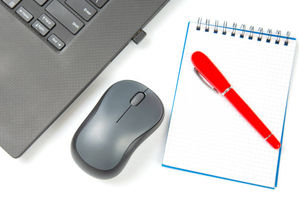 istock Computer wireless mouse, pen and notepad next to a laptop on a white table close-up. Items for office work 1413627501