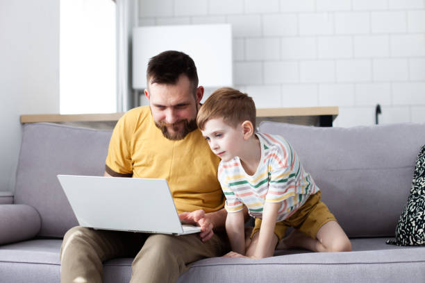 Father and his little son looking something interesting at computer screen using laptop at home. stock photo