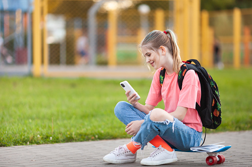 Teen schoolgirl with backpack sitting on skateboard in a school yard. Blond girl with colored hair strands looking in smartphone. Back to school concept. Selective focus.