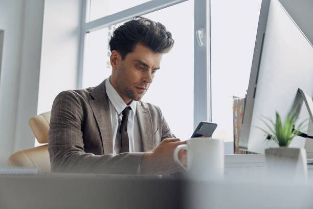 istock Concentrated young man using smart phone while sitting at his working place in office 1413629987