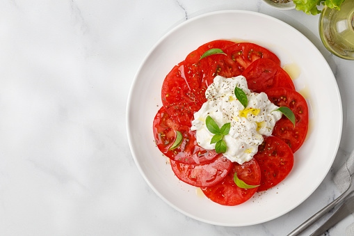 Strachatella salad with circle of sliced tomatoes, oil and basil on a white plate. Top view, copy space. Italian food