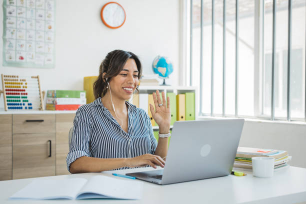 Elementary school teacher Teacher at classroom sitting at desk and using laptop. She is talking with students on video call. online tutors stock pictures, royalty-free photos & images
