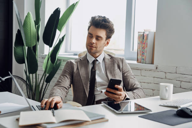 istock Confident young man using laptop and holding smart phone while sitting in 1413631045
