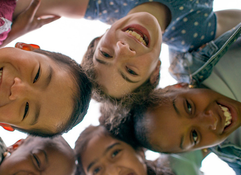 A small group of 5 school aged children press their heads together as they look down to the ground.  They are each dressed casually and smiling at the camera below.