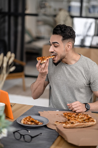 Eating at home. Happy young bearded man biting pizza looking interestedly at tablet screen sitting at table at home