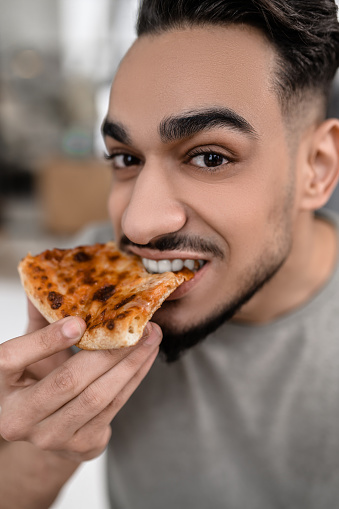 Pleasure. Close-up face of young dark-haired man looking at camera eating pizza holding piece near mouth