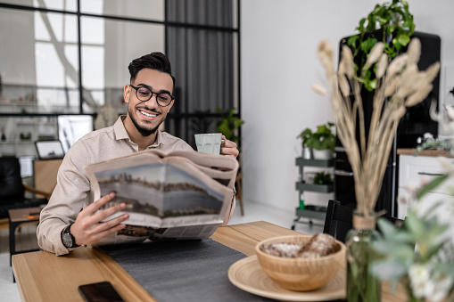 Morning news. Cheerful young bearded man in glasses sitting with coffee reading interested newspaper sitting at table in morning