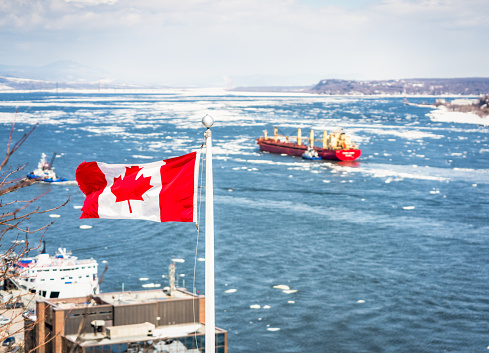 A Canadian flag flying high over Quebec City in early springtime, with ships in the St. Lawrence Seaway defocused in the background.